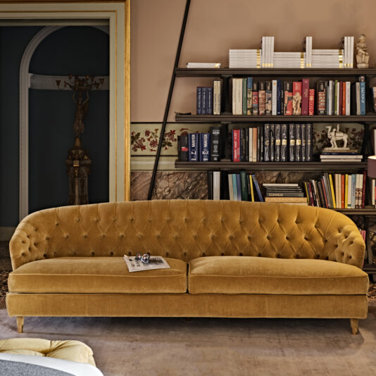A lavish living room features the Large Button Upholstered Velvet Sofa in mustard yellow, centered in the foreground. Behind the sofa is a tall bookshelf filled with books and decorative items. The walls are adorned with classic molding and artwork. A newspaper and glasses rest on the sofa.