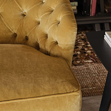 Large Button Upholstered Velvet Sofa Close-up of a mustard yellow tufted velvet armchair next to a dark wooden side table. In the background, there's a glimpse of a bookshelf with several books and a patterned rug on the floor. The scene is complemented by the Large Button Upholstered Velvet Sofa nearby, creating an inviting and cozy reading nook.