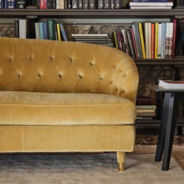 Large Button Upholstered Velvet Sofa A Large Button Upholstered Velvet Sofa in mustard yellow is positioned in front of a bookshelf filled with an assortment of books. To the right of the sofa is a black wooden side table with a stack of books on top. The scene is cozy and inviting, perfect for a charming reading nook.