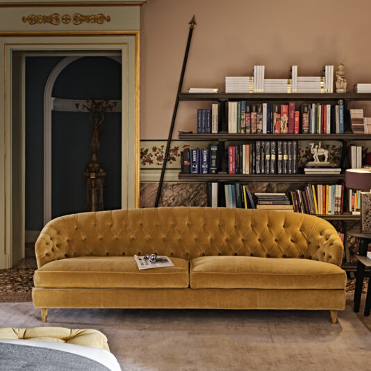 A cozy living room features a Large Button Upholstered Velvet Sofa in mustard yellow prominently positioned at the center. In front of it stands a dark wooden bookshelf filled with books and decorative items. The beige walls are adorned with ornate wooden trim, and a doorway is visible in the background.