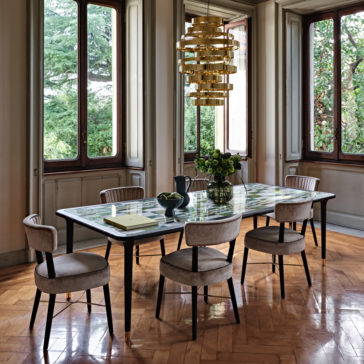 A well-lit dining room with large windows revealing lush greenery outside. The room features a rectangular table adorned with a colorful checkered top and six upholstered chairs. Centrally located above the table is the Large Contemporary Chandelier, showcasing stunning gold cylindrical designs. A vase of flowers adds a touch of nature at the center of the table.