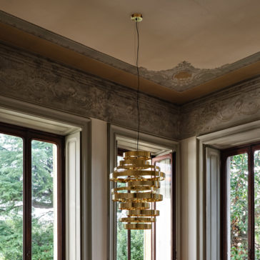A Large Contemporary Chandelier, featuring a gold-colored spiral design, hangs elegantly from the ceiling in a room adorned with ornate molding and large windows that offer a view of trees outside. The space exudes sophisticated and classical charm.