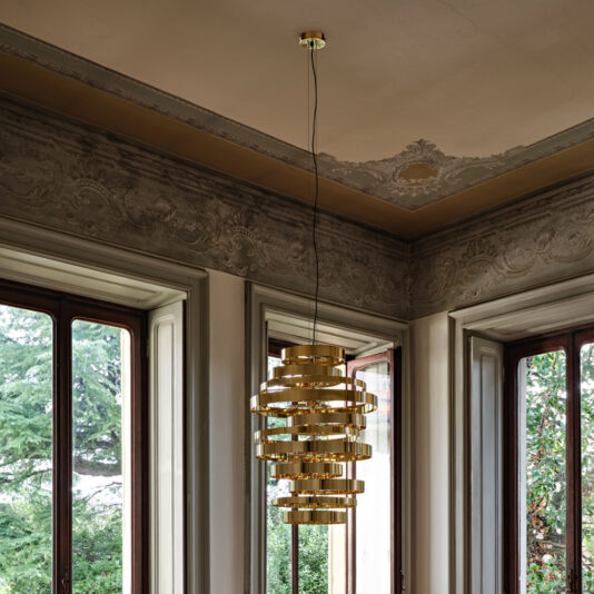 Large Contemporary Chandelier A Large Contemporary Chandelier, featuring a gold-colored spiral design, hangs elegantly from the ceiling in a room adorned with ornate molding and large windows that offer a view of trees outside. The space exudes sophisticated and classical charm.