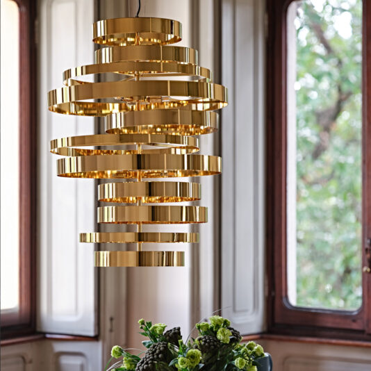 Large Contemporary Chandelier A large Contemporary Chandelier with a spiral design and gold finish illuminates a room featuring expansive windows. The light-colored walls harmonize with the bouquet of green flowers arranged in a clear vase on the table below. Through the windows, trees infuse nature into the elegantly lit space.