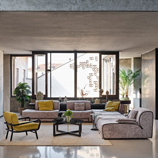 A modern living room with large windows letting in natural light features a Large Contemporary Modular Corner Sofa as the centerpiece, adorned with yellow cushions. The space is further complemented by a matching yellow armchair and a beige chaise lounge. Black coffee tables are scattered throughout the room, while plants near the windows and framed photos on the wall enhance its charm.