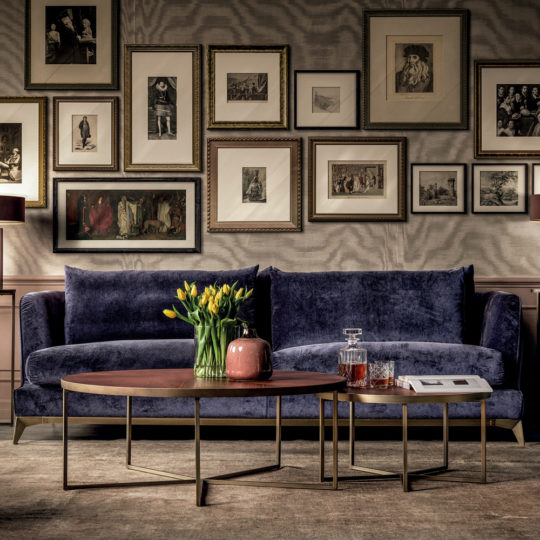 A sophisticated living room featuring a large luxury contemporary sofa in dark blue velvet, adorned with a vase of yellow tulips on a modern round coffee table. The wall behind the stylish sofa is decorated with an eclectic gallery of framed art and photographs. Two side tables hold a decanter and some books.