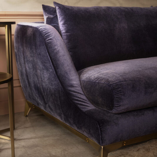 A close-up view of the Large Luxury Contemporary Sofa showcases its deep purple velvet with a plush cushion and sleek gold legs. Positioned in a cozy, stylish room featuring neutral-toned walls and complemented by a small round side table, the lighting enhances the fabric's rich, soft texture.