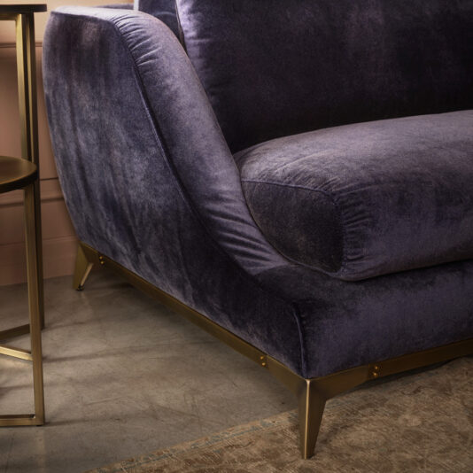 Close-up view of the corner of the Large Luxury Contemporary Sofa in dark purple velvet with a gold metal base, paired with a gold side table on a concrete floor and a beige carpet. The background includes a section of a pink wall.