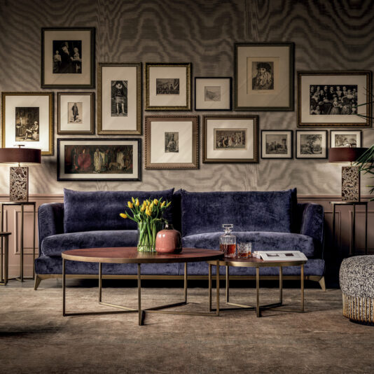 A stylish living room featuring the Large Luxury Contemporary Sofa in navy blue velvet, adorned with yellow tulips in a pink vase placed on a set of modern nested coffee tables. The wall behind is decorated with an eclectic gallery of framed photos and art. Warm, ambient lighting enhances the cozy, luxury scene.
