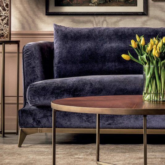 A cozy living room showcases the Large Luxury Contemporary Sofa in dark blue velvet, paired with a round wooden coffee table on a brass base and yellow tulips. A framed artwork partly adorns the wall above, while a light beige rug covers the floor.