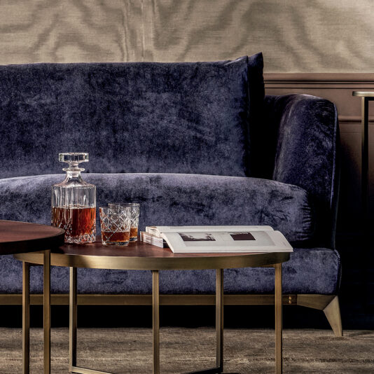 A Large Luxury Contemporary Sofa in dark blue velvet sits against a beige wall. In front, there is a wooden coffee table with a glass decanter of amber liquid and a matching glass. Adjacent is a smaller table with a white book or newspaper on top. Both tables have brass frames, adding to the contemporary feel.