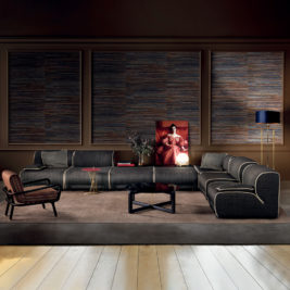 A modern living room features the Large Modular Corner Sofa in dark gray with light piping, there's occasional seating in the form of a wooden armchair, a red side table, and a black coffee table on a beige rug. The wall behind has three sections with textured dark blue and brown panels and a framed artwork.