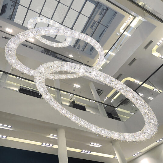 The Luxury Oval Crystal Chandelier, featuring large, interlocking circular rings illuminated with numerous small lights, hangs from the ceiling of an atrium with glass railings and multiple levels. It creates an elegant and contemporary atmosphere.