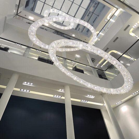 A modern, multi-story interior space features the Luxury Oval Crystal Chandelier, a sophisticated fixture with two intersecting rings. The high ceilings reveal multiple floors adorned with sleek glass railings, all brilliantly illuminated by bright lighting that enhances the area's elegance.
