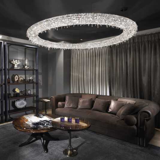 A modern living room showcasing the Luxury Oval Crystal Chandelier, a dark brown plush couch adorned with several pillows, a rounded wood coffee table, and a tall shelving unit filled with decorative items. The space is dimly lit and features floor-length curtains behind the couch.