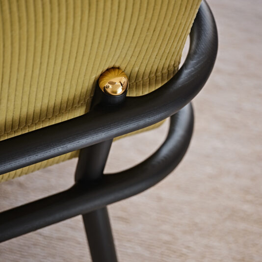 Close-up of a backrest on the Low Mid Century Style Arm Chair, featuring vertical ridged olive green upholstery. The back is secured by a golden knob attached to an elegant, smooth black tubular metal frame. The surface beneath the chair seems to be a light-colored fabric or carpet.