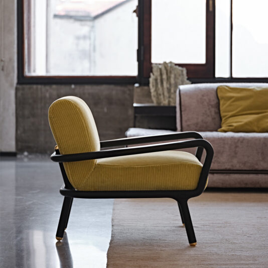 A modern interior with the Low Mid Century Style Arm Chair upholstered in mustard, featuring black wooden armrests and legs, placed on a light-colored rug. In the background, a grey sofa with a yellow cushion sits near large windows that offer partial views of an outdoor scene.