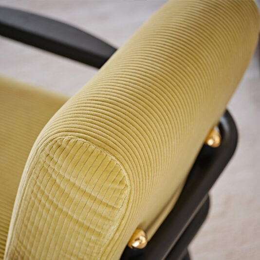 Close-up of the armrest of the Low Mid Century Style Arm Chair, showcasing its yellow corduroy fabric with horizontal ridges. The chair, set in a black frame and highlighted with gold-colored fasteners, exudes a stylish and contemporary appeal.