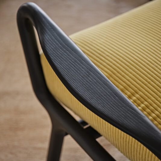 Close-up of the Low Mid-Century Style Arm Chair, featuring a black wooden frame and a mustard-yellow ribbed cushion. The chair's sleek design and fabric texture are prominently displayed, with a slightly blurred background emphasizing the chair's intricate details.