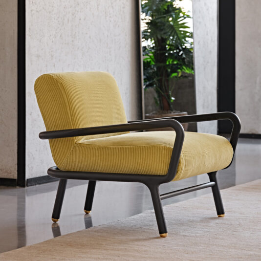 A Low Mid Century Style Arm Chair with yellow upholstery and a black metal frame sits on a light-colored rug in a modern room. The chair features a rounded backrest and armrests, with greenery visible by the large window. The polished concrete floor adds to the sleek aesthetic.