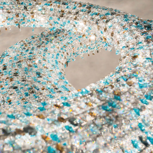 Luxury Coloured Crystal Chandelier A close-up photo of an intricate sculpture made up of hundreds of shimmering blue, white, and clear beads interwoven in a fluid, swirling pattern. Sunlight reflects off the beads, creating a sparkling effect reminiscent of the Luxury Coloured Crystal Chandelier. The shapes suggest a dynamic, flowing motion.
