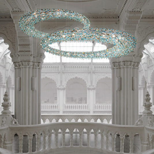 Luxury Coloured Crystal Chandelier A grand hall features an ornate, white architectural design with tall columns and decorative arches. Hanging from the ceiling are two Luxury Coloured Crystal Chandeliers, their elegant circular forms and exquisite crystals creating a sophisticated and luxurious atmosphere.