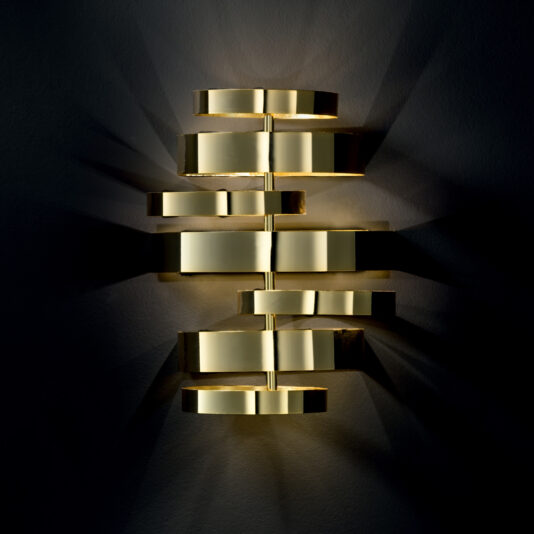 The Luxury Contemporary Wall Light, crafted from polished gold-colored metal, boasts a modern design with overlapping horizontal rectangular panels arranged in a staggered vertical pattern, creating a soft, warm glow against a dark background.