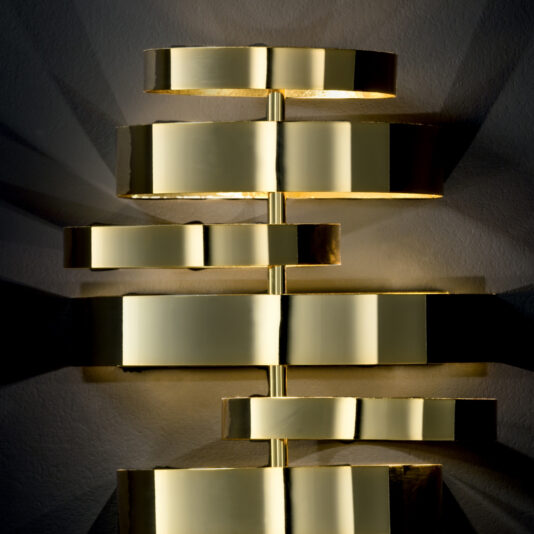 Luxury Contemporary Wall Light The Luxury Contemporary Wall Light is a sophisticated fixture that boasts several horizontally stacked, gold-colored geometric shapes arranged at various angles. These golden surfaces reflect light beautifully, casting a warm glow and intricate shadows on the wall behind.