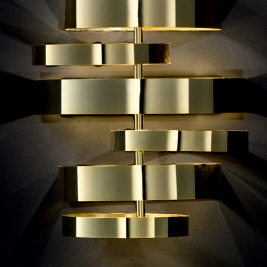 Luxury Contemporary Wall Light A close-up photograph of the Luxury Contemporary Wall Light showcases its series of horizontal, metallic gold bands and shapes. The light casts intricate, symmetrical shadows on the wall behind it, creating a striking visual effect.