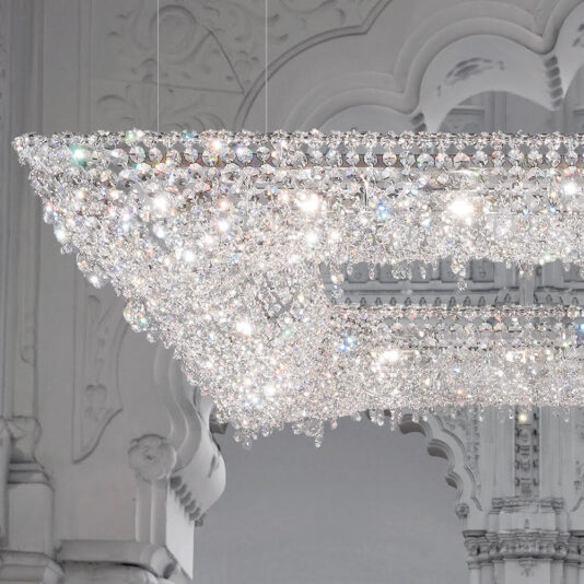 Luxury High End Crystal Chandelier A Luxury High End Crystal Chandelier hangs from an ornate ceiling adorned with detailed molding. This exquisite chandelier, featuring numerous sparkling crystals, disperses light in multiple directions, creating a dazzling effect that beautifully complements the elegant architectural details in the background.