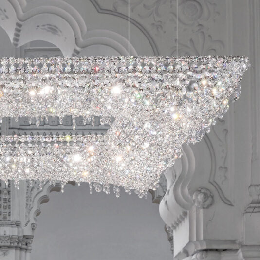 Luxury High End Crystal Chandelier The Luxury High End Crystal Chandelier, a large and rectangular fixture adorned with intricate details and numerous sparkling crystals, is suspended from the ceiling. Its illumination creates a dazzling display of light and reflections on the ornate ceiling and wall behind it.
