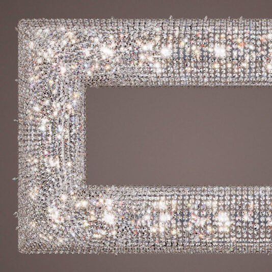 Luxury High End Crystal Chandelier A close-up image of the Luxury High End Crystal Chandelier reveals its rectangular mirror frame adorned with numerous small, glittering crystals, evoking opulence. The design exudes elegance with its dazzling and reflective surface, set against a neutral gray background.