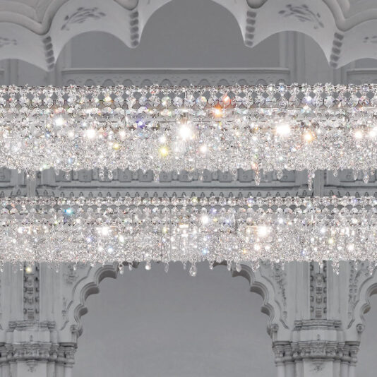 Luxury High End Crystal Chandelier A grand, Luxury High End Crystal Chandelier with multiple layers of sparkling crystals is suspended in front of an ornate, white architectural background featuring decorative arches and intricate detailing. The chandelier reflects light beautifully, creating a stunning, glittering effect.