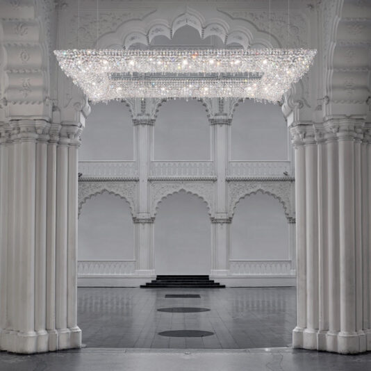 Luxury High End Crystal Chandelier The spacious hall, decorated with ornate arches and columns featuring intricate carvings, is illuminated by the Luxury High End Crystal Chandelier. This elegant fixture, adorned with numerous crystals, harmonizes perfectly with the palatial architectural style and neutral tones of the setting, epitomizing ultimate luxury.