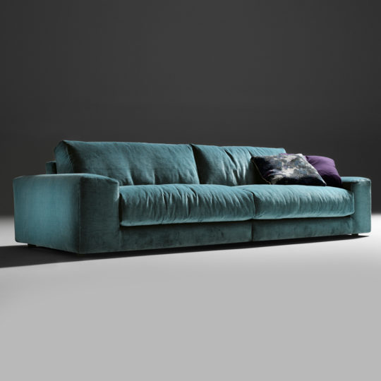 The Luxury Large Velvet Modular Sofa in modern teal is set against a dark background, featuring plump cushions and armrests on both sides. This sophisticated sofa is complemented by two decorative pillows, including one in dark blue with a subtle pattern, enhancing its sleek and elegant design.