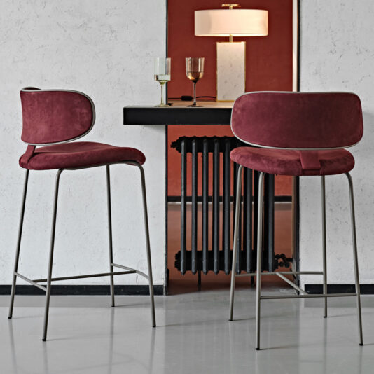Luxury Mid Century Style Bar Stool Two Luxury Mid Century Style Bar Stools featuring red cushions and sleek metal legs are positioned by a minimalistic bar counter. Atop the counter, two wine glasses sit beside a modern table lamp adorned with a beige lampshade. The textured wall behind the counter enhances the contemporary ambiance.