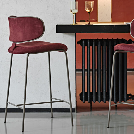 Luxury Mid Century Style Bar Stool A sophisticated bar area boasts the Luxury Mid Century Style Bar Stools, showcasing plush burgundy suede seats and backs anchored by sleek metal frames. A chic black bar counter stands against a lightly textured wall, topped with an assortment of glasses for a refined touch. Beneath the counter, a black radiator is subtly visible, completing the elegant design.