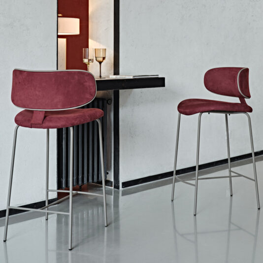 Luxury Mid Century Style Bar Stool A modern bar interior showcases two Luxury Mid Century Style Bar Stools, each featuring maroon seats with sleek silver legs. This stylish setup is paired with a small, dark bar counter adorned with a wine glass and tumbler, seamlessly mounted on the wall. The entire ensemble is accented by white and black decor, creating a minimalist design with clean lines against a neutral background.