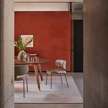 A stylish dining area with a circular wooden table, light grey cushioned Luxury Mid Century Style Dining Chairs, and a vase of green and white flowers as the centerpiece. The room features a rich red accent wall with minimalist floral artwork, framed by rough concrete walls.