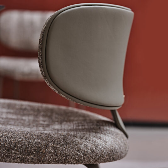 Luxury Mid Century Style Dining Chair Close-up of the Luxury Mid Century Style Dining Chair featuring a curved backrest upholstered in smooth, light brown leather and a padded seat covered in textured fabric. Its mid-century design stands out against a red wall with the blurred outline of another similar chair, creating a contemporary setting.
