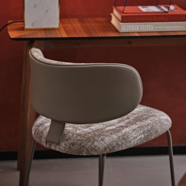 A Luxury Mid Century Style Dining Chair with a curved backrest upholstered in brown leather and a textured fabric seat is placed near a wooden desk. On the desk are books, including one titled 