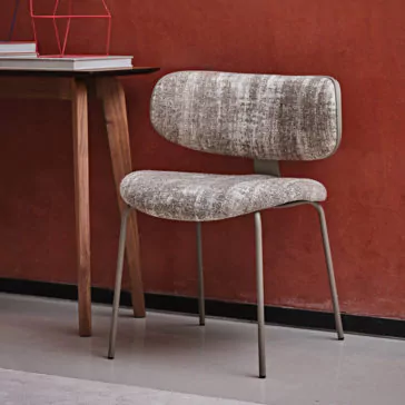 A Luxury Mid Century Style Dining Chair, featuring a textured gray and beige fabric seat and backrest supported by thin metal legs, is placed beside a wooden desk with books on it, against a red wall.