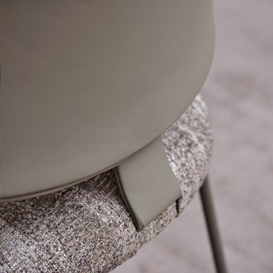Luxury Mid Century Style Dining Chair Close-up of the Luxury Mid Century Style Dining Chair, revealing a backrest elegantly covered in smooth gray leather and a seat upholstered in textured beige fabric. A sophisticated leather detail seamlessly connects the backrest to the seat cushion, while the partially visible legs provide sturdy support for the cushioned sections.