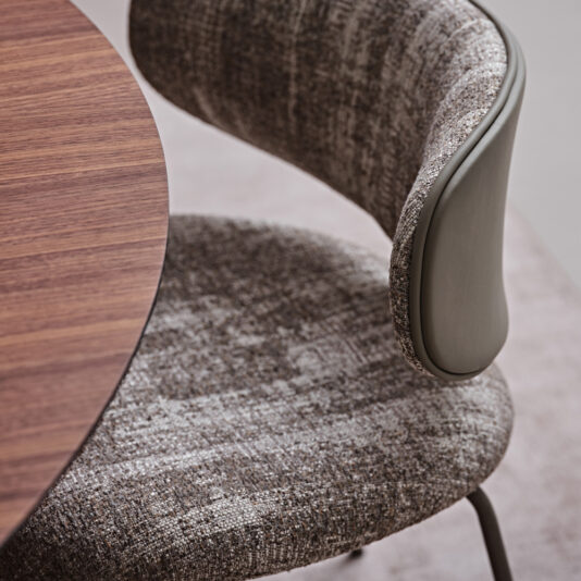 Luxury Mid Century Style Dining Chair A close-up view showcases the Luxury Mid Century Style Dining Chair alongside part of a round wooden table. This chair boasts textured, patterned upholstery in shades of gray and brown, complemented by a sleek, curved backrest with a smooth exterior. The setting appears to be a contemporary interior space with mid-century style influences.