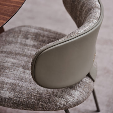 Close-up of the Luxury Mid Century Style Dining Chair, showcasing its modern upholstered seat and backrest with textured fabric. The chair features a sleek, curved design, with the back wrapped in smooth, leather-like material and supported by slim metal legs. This exquisite piece is positioned next to a wooden table.