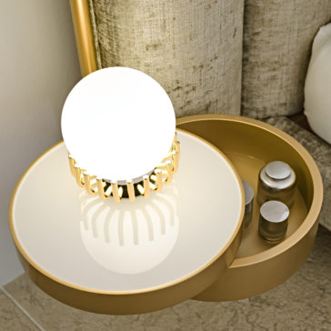 A contemporary circular bedside table with a sleek white surface and an integrated orb light, accented by a gold border. Next to it, a small compartment houses several labeled jars. This setup is complemented by the Luxury Modern Bed With Wide Headboard, featuring a textured beige finish.