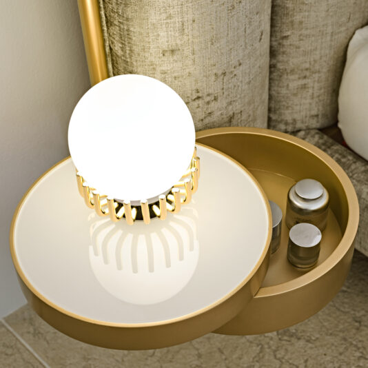 Luxury Modern Bed With Wide Headboard A contemporary circular bedside table with a sleek white surface and an integrated orb light, accented by a gold border. Next to it, a small compartment houses several labeled jars. This setup is complemented by the Luxury Modern Bed With Wide Headboard, featuring a textured beige finish.