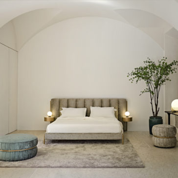 A minimalist bedroom features the Luxury Modern Bed With Wide Headboard, upholstered in beige and paired with white bedding. Flanked by round bedside tables with dome lamps, the space includes a tall green plant, a round ottoman, and a smaller stool in soft hues on a textured rug.