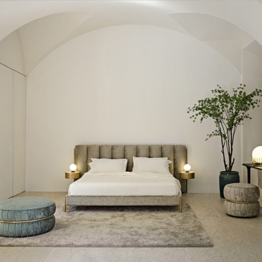 Luxury Modern Bed With Wide Headboard A minimalist bedroom features the Luxury Modern Bed With Wide Headboard, upholstered in beige and paired with white bedding. Flanked by round bedside tables with dome lamps, the space includes a tall green plant, a round ottoman, and a smaller stool in soft hues on a textured rug.