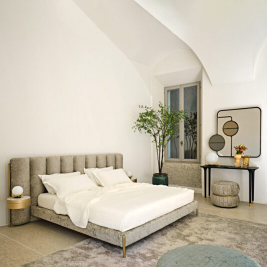 Luxury Modern Bed With Wide Headboard A contemporary bedroom featuring a Luxury Modern Bed With Wide Headboard, adorned with white bedding. A small tree in a pot stands beside the window. A round table with lamps and decor items sits nearby, along with a mirror and an ottoman. The room boasts soft lighting and minimalistic decor.